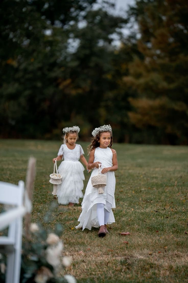 Two flower girls in white dresses and floral crowns walking down a garden aisle
