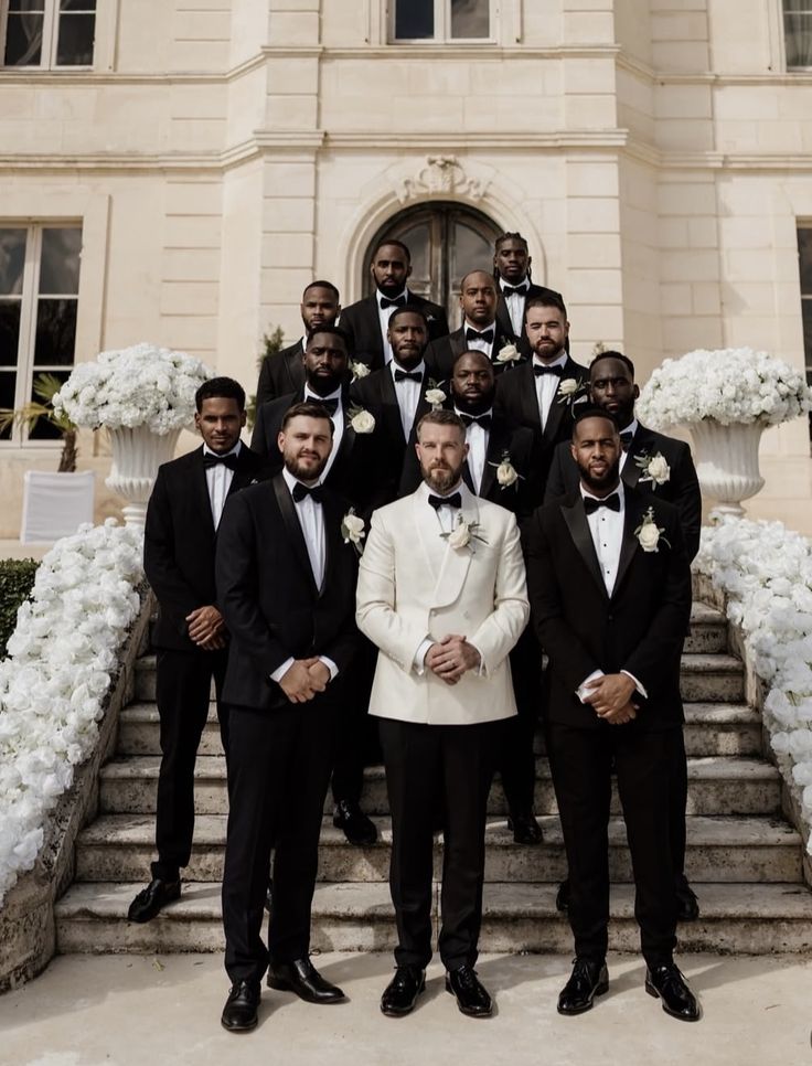 Groom in an ivory tuxedo standing with groomsmen in black tuxedos on stone steps surrounded by white flowers